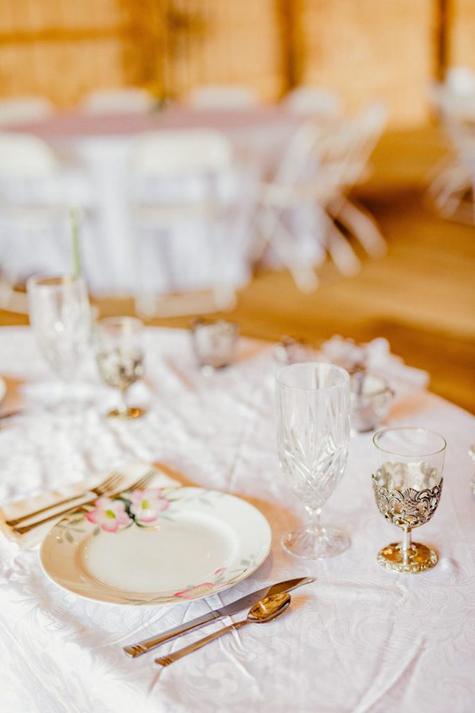 Delicate china, glassware, and silverware sit upon pristine white tablecloth inside the reception barn.